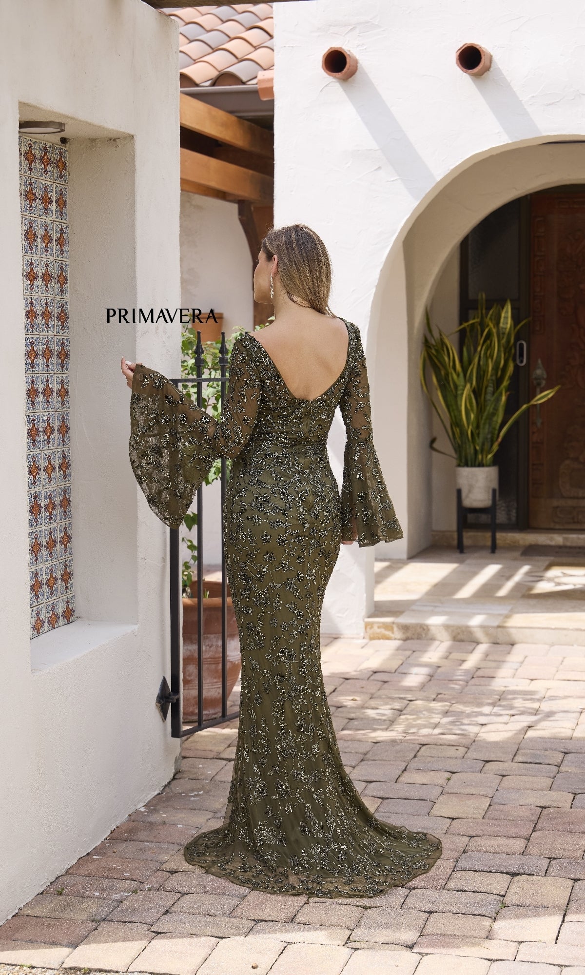 A woman wearing the Primavera 12185 long formal dress in olive green, featuring beads, bell sleeves, and flared cuffs, stands in a sunlit courtyard facing away from the camera.
